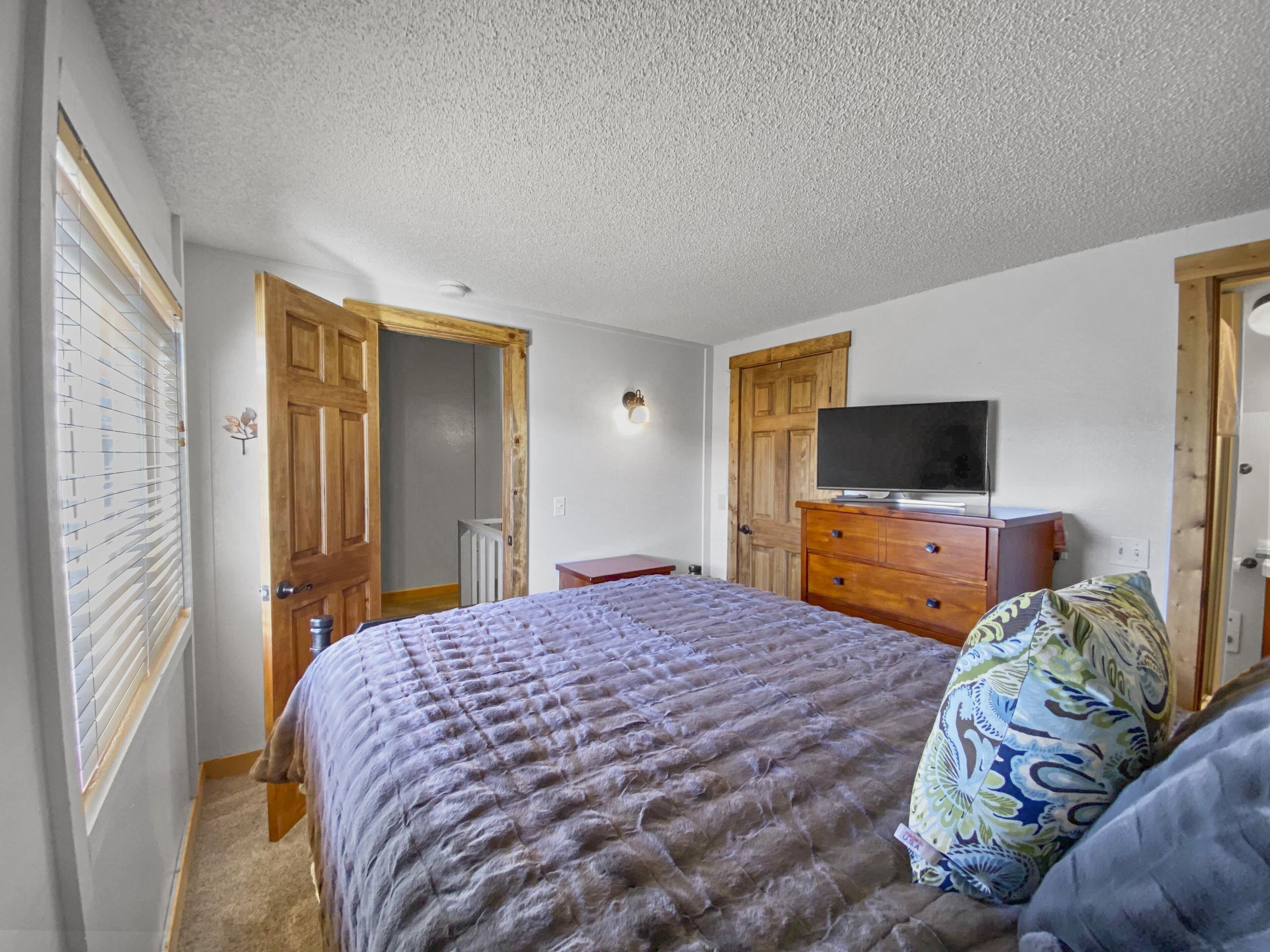 A bedroom with a bed, a television on a wooden dresser, and a door leading to a hallway.