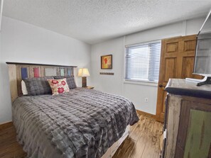 A bedroom with a bed, a wooden headboard, a nightstand with a lamp, a window with blinds, a door, and a television on a wooden cabinet. - Powder Ridge 209 4 Bedroom Condo by RedAwning (Breckenridge)