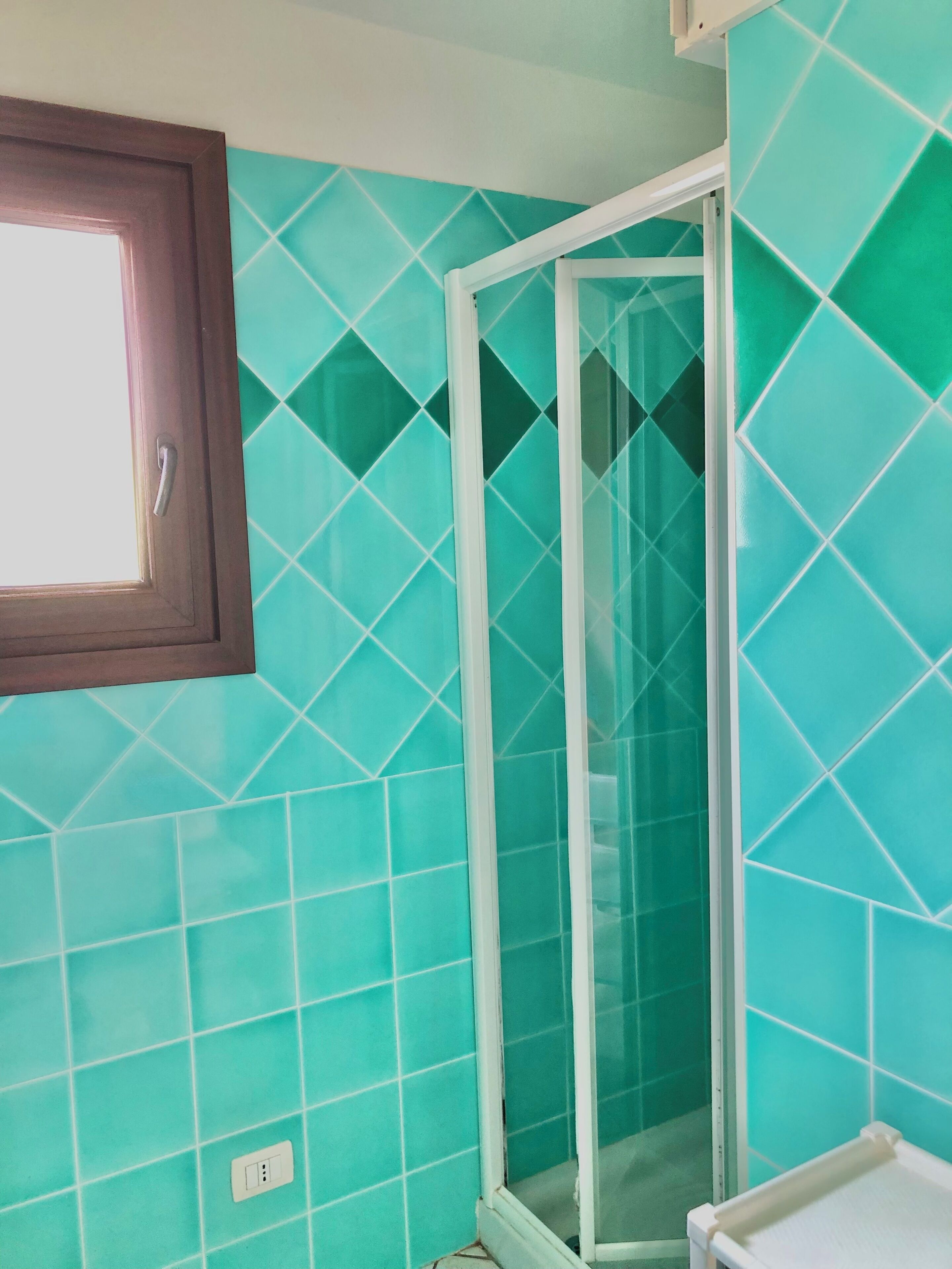A bathroom with teal tiles, a glass shower enclosure, and a wooden-framed window.