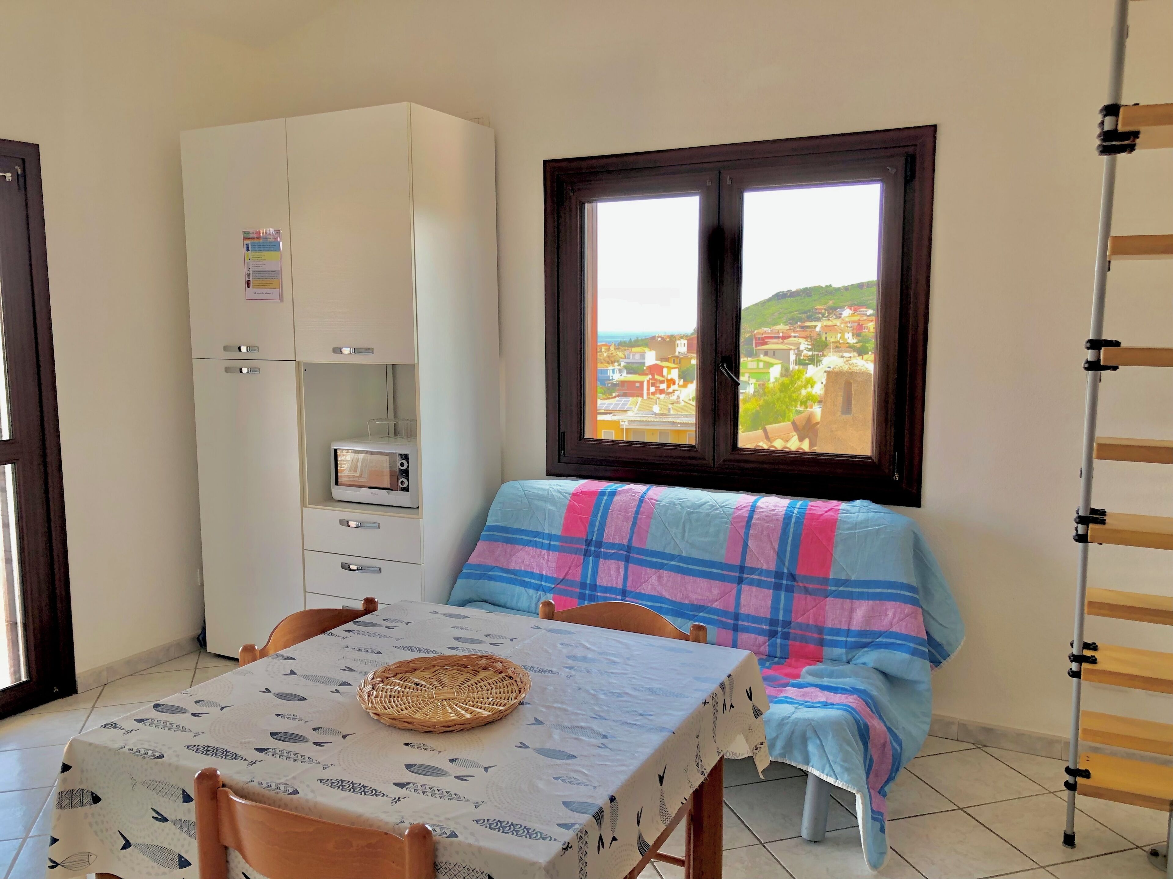 Una habitación pequeña con una mesa de comedor, un sofá con una manta escocesa, un armario blanco con un microondas y una ventana con vistas a edificios.