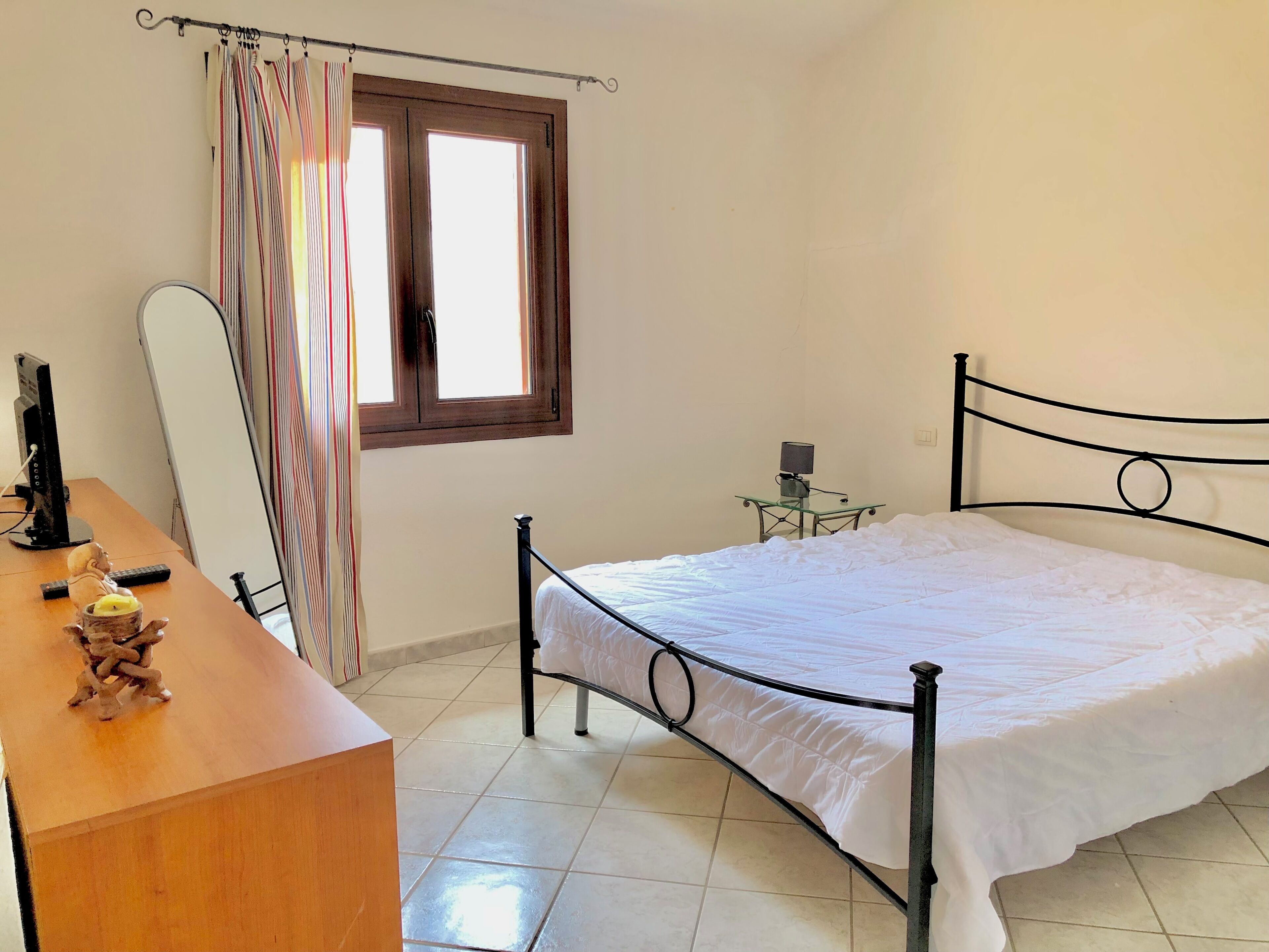 A bedroom with a bed, a desk with a computer, a small table with a lamp, and a window with curtains.