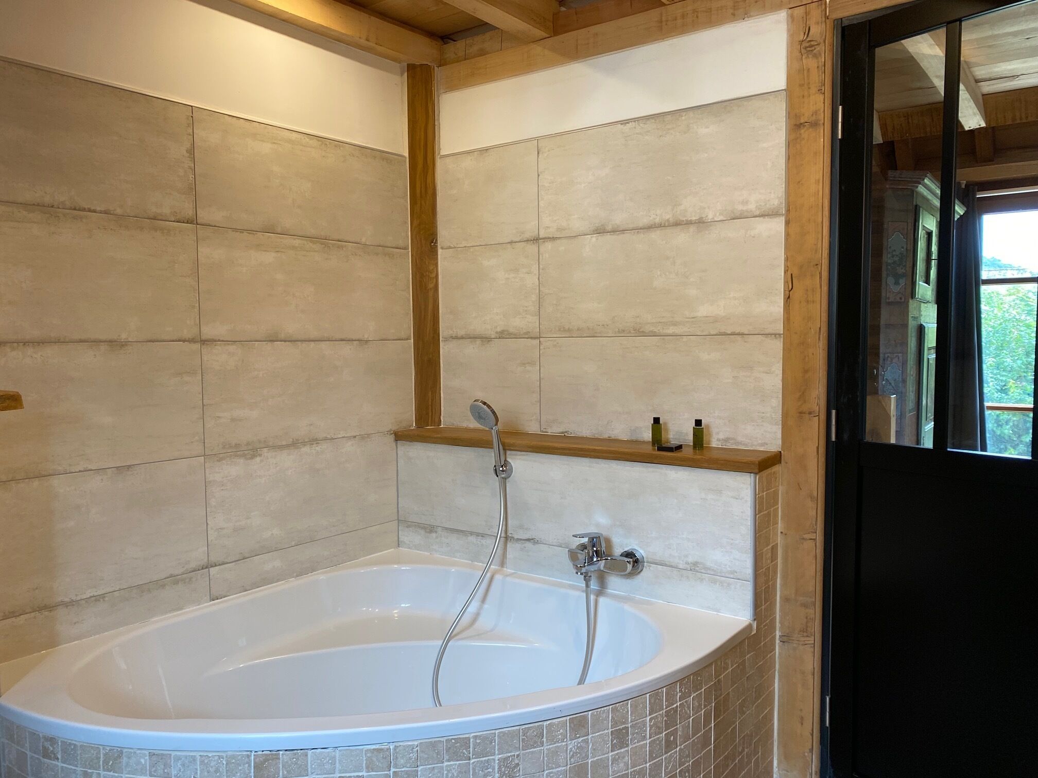 A modern bathroom with a freestanding bathtub, a wooden shelf above it, and a tiled wall.