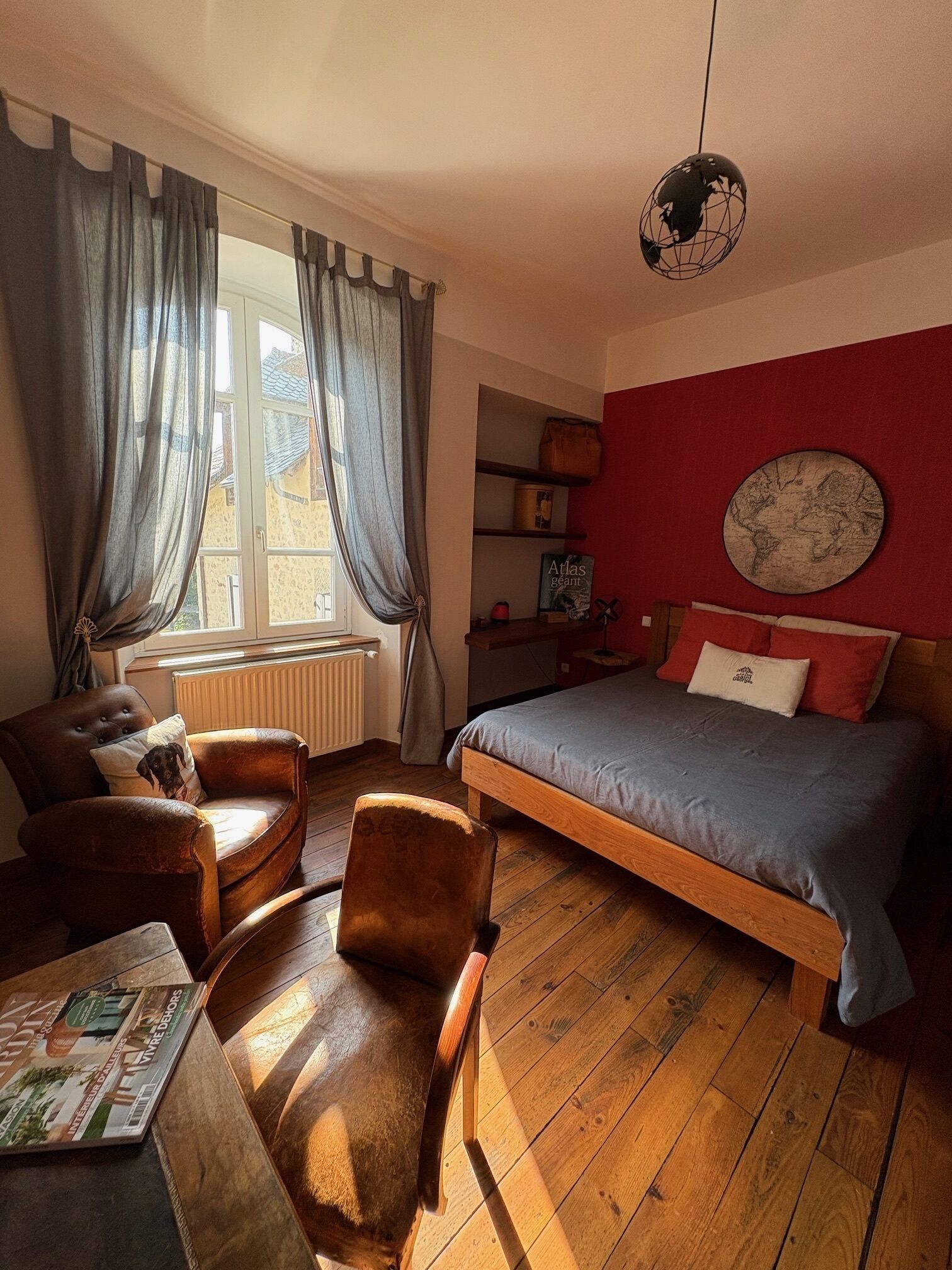 Une chambre avec un plancher en bois, un lit recouvert d’une courtepointe bleue, un fauteuil en cuir marron, une table de chevet en bois, un globe suspendu au plafond et une fenêtre avec des rideaux.