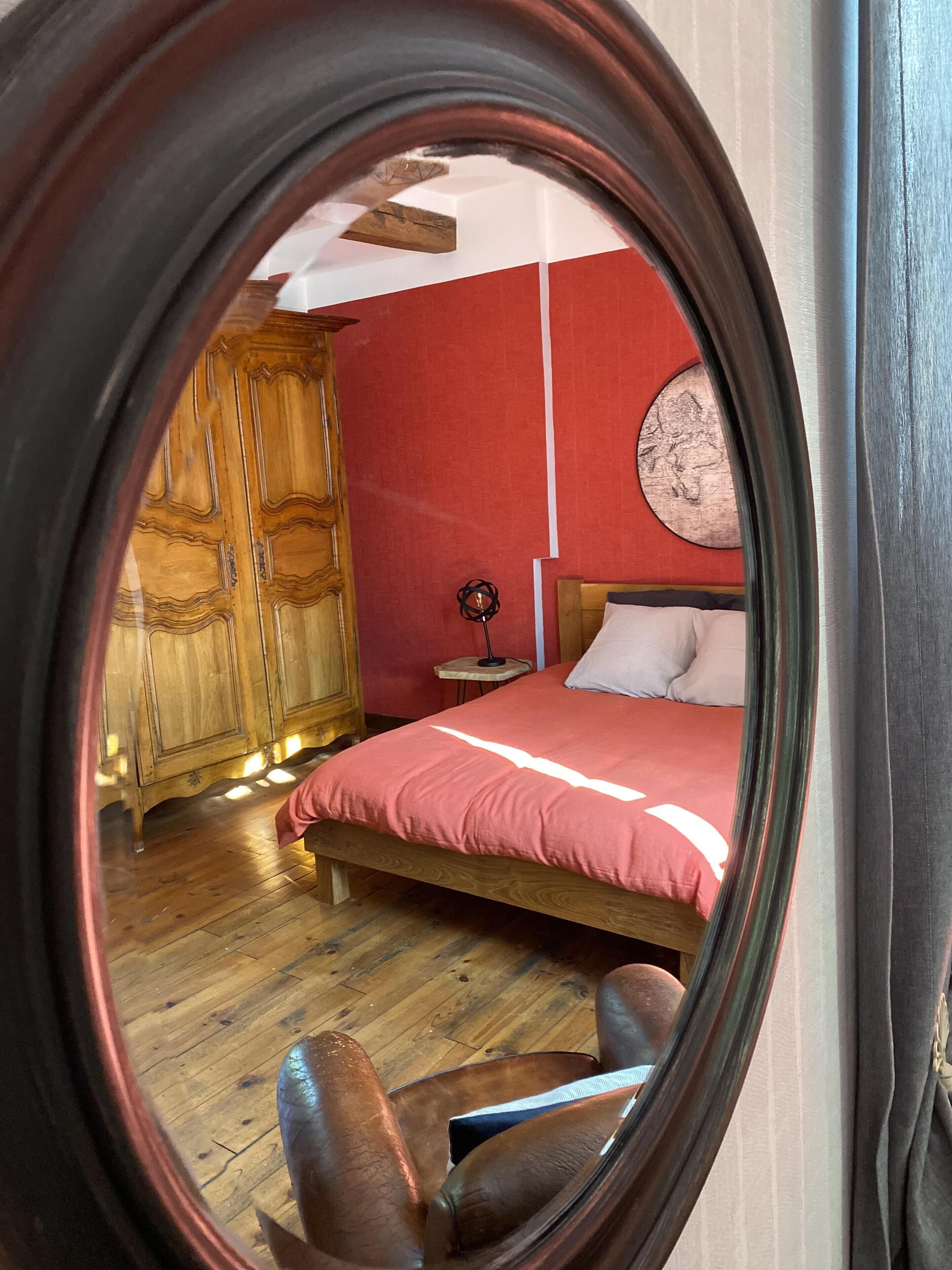 A bedroom with a wooden wardrobe, a round mirror, a bed with a red bedspread, and a small round table with a lamp.