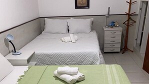 Une chambre d’hôtel avec deux lits, l’un recouvert d’une couvre-lit verte et l’autre d’une couvre-lit blanche. On y trouve une table de chevet avec une lampe, une commode et un porte-manteau.