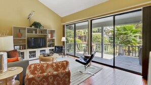 Un salon spacieux doté d’une grande télévision, de sièges confortables, d’un balcon offrant une vue sur la verdure et d’un design moderne.