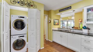 Une cuisine avec des meubles blancs, un évier et un plan de travail. On y trouve deux machines à laver superposées et une décoration représentant un vélo accrochée au mur.