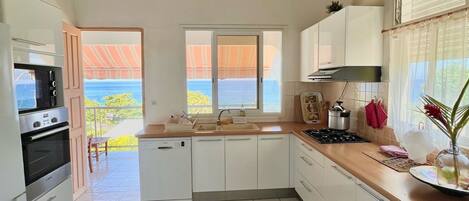 A modern kitchen with white cabinets, a built-in oven, and a view of the sea through the open door.