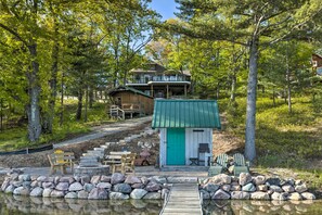 A lakeside cabin with a dock leading to a small docked boat, surrounded by trees and a wooden deck with outdoor seating.