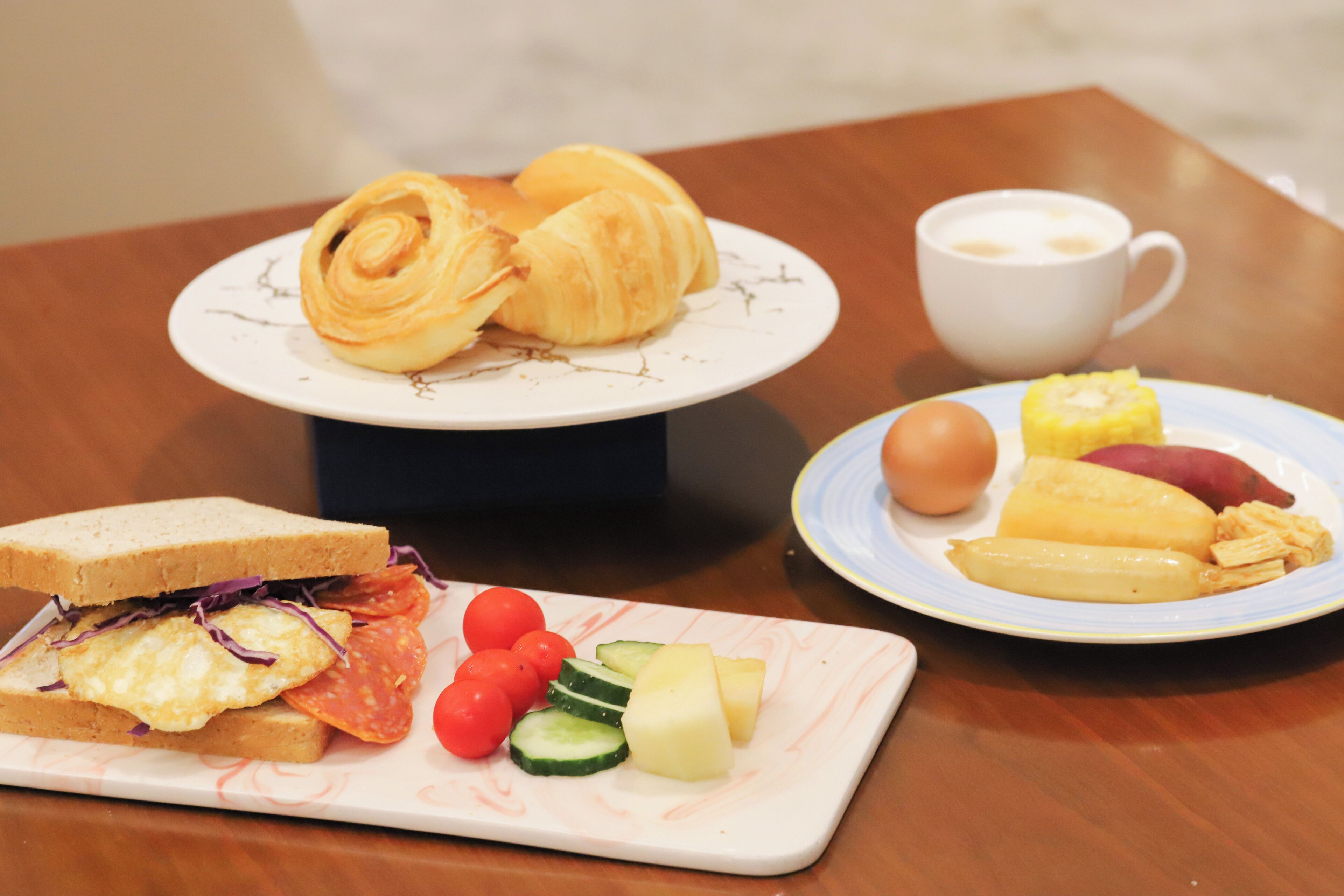 A breakfast setting with croissants, a sandwich, eggs, tomatoes, cucumbers, and a cup of coffee on a table.