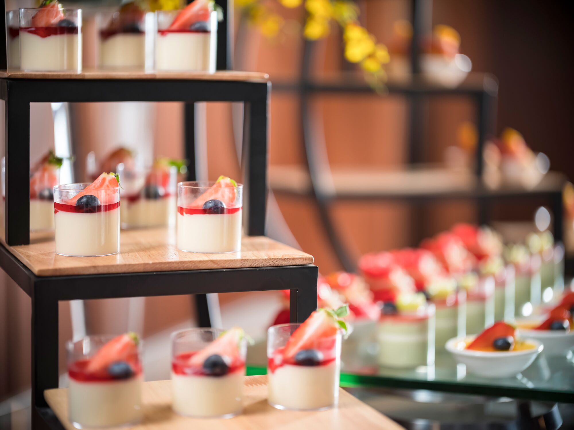 A dessert display with multiple glass servings on a wooden stand, each topped with a strawberry and a blueberry.