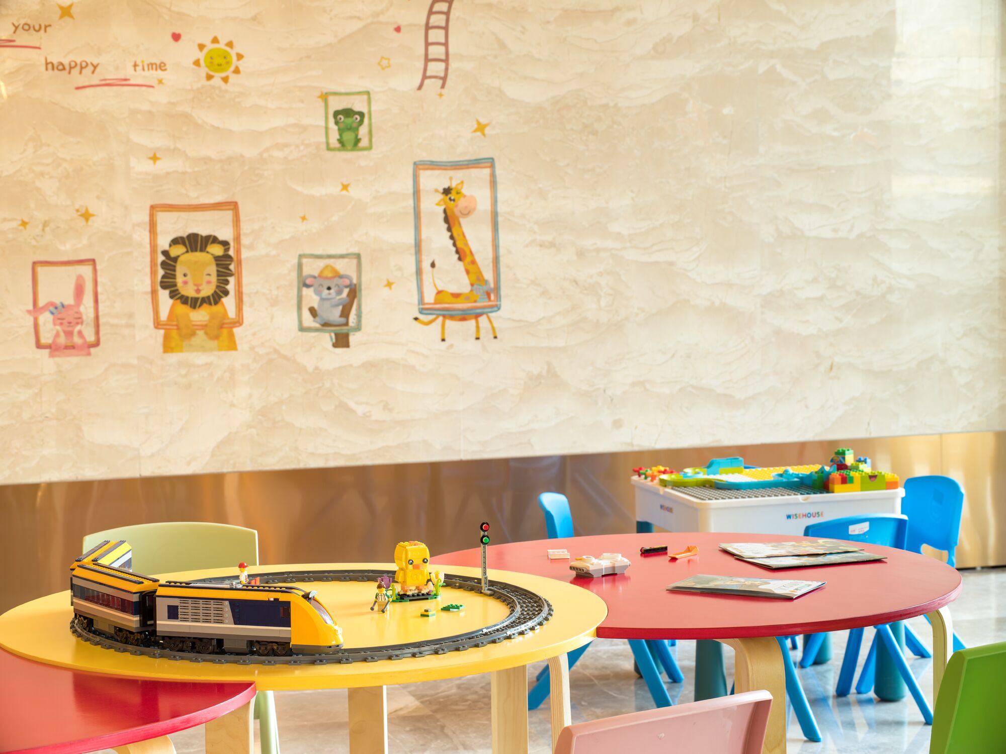 Área de recreação infantil com um conjunto de trens em uma mesa amarela, cadeiras coloridas e uma parede decorada com imagens de animais e uma escada.