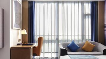 Habitación de hotel con escritorio, silla, cama y ventanas amplias con cortinas.