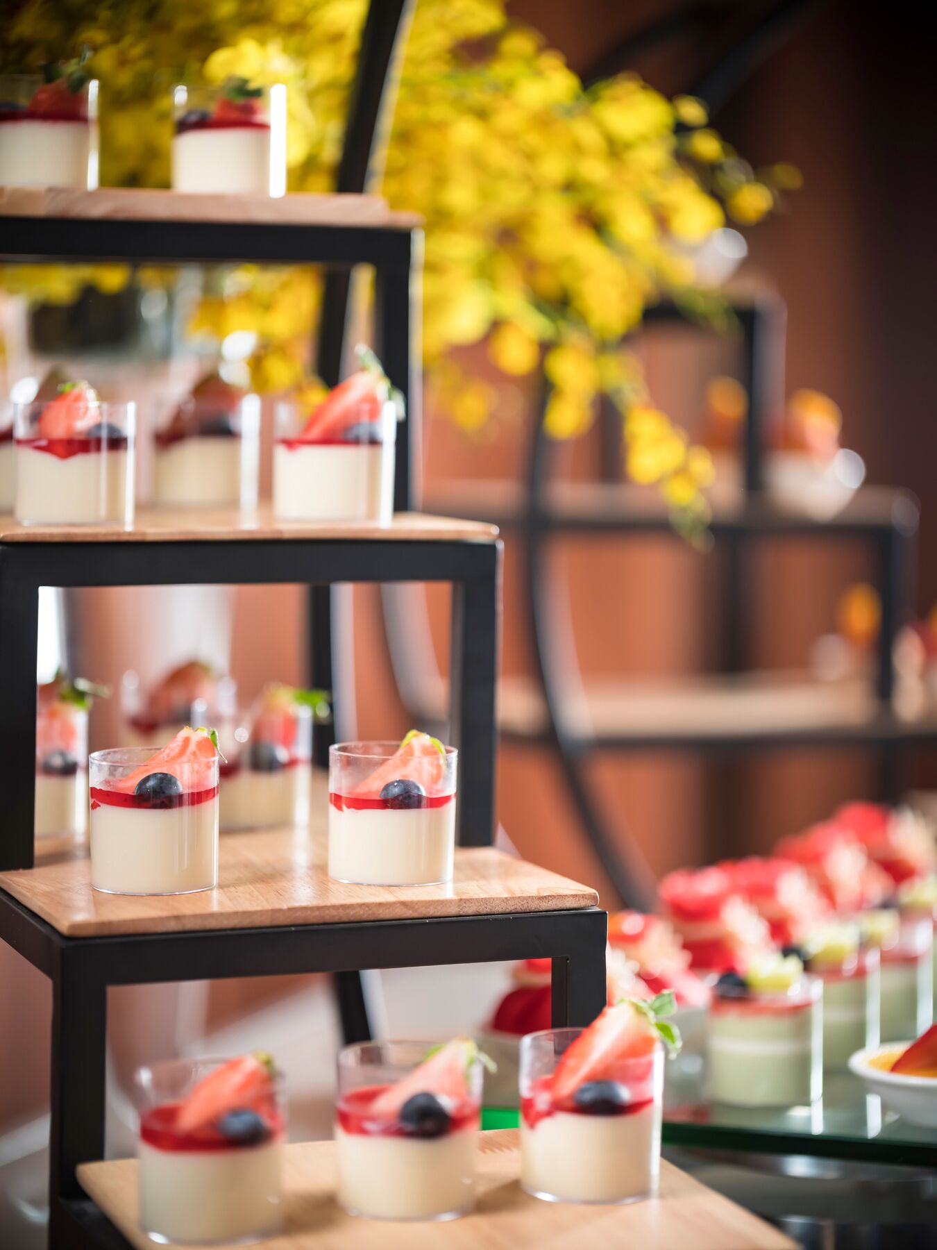 Una exhibición de postres con varios niveles de copas de vidrio que contienen postres en capas, sobre una mesa de madera con un fondo borroso de flores amarillas.