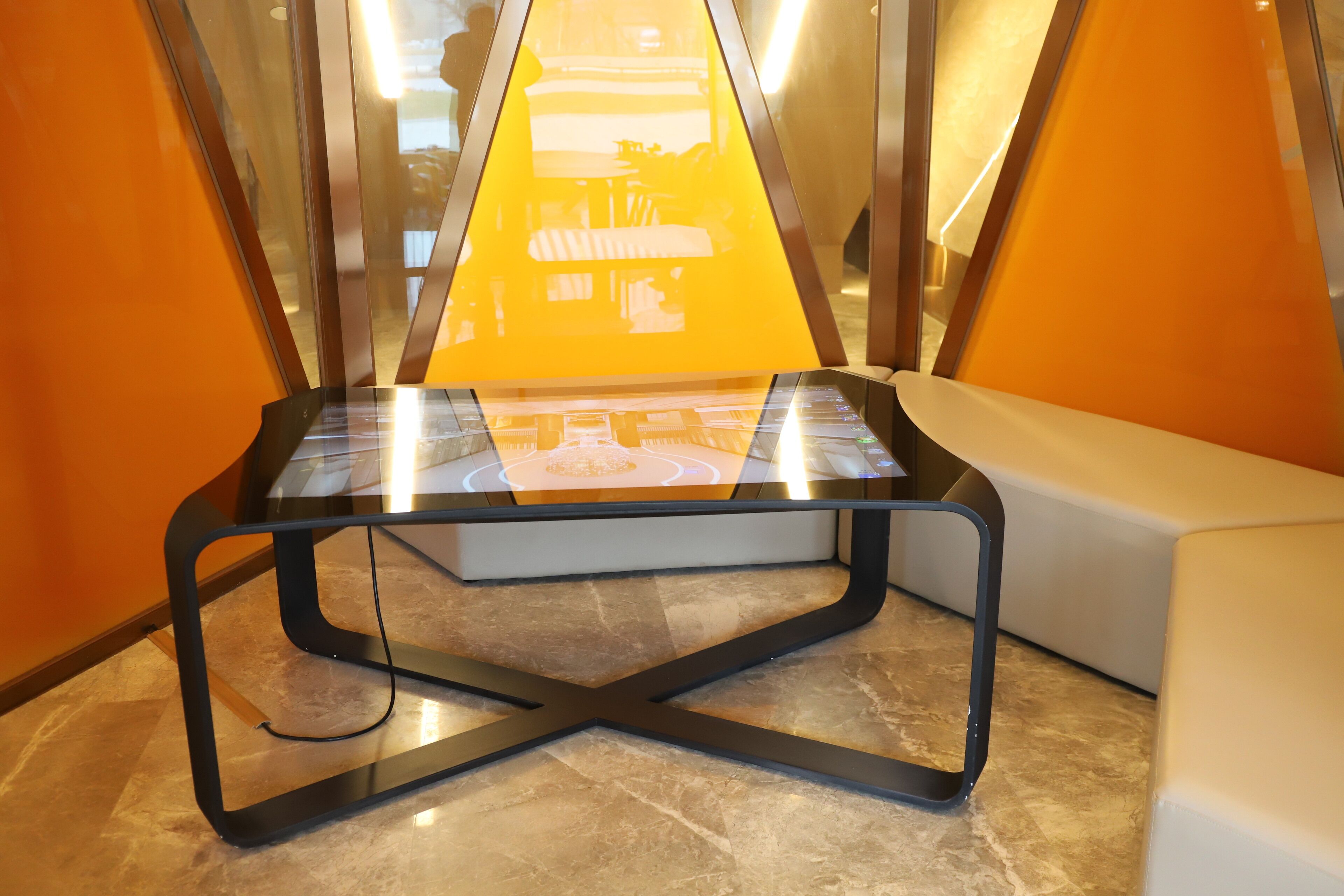 A modern, glass-top table with a black metal frame, positioned in a room with orange walls and a marble floor.