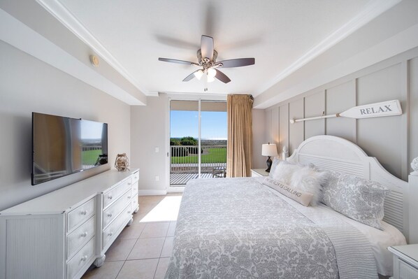 A bedroom with a large bed, a ceiling fan, a TV, a dresser, and a view of the outdoors.
