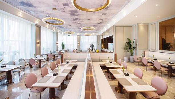 Un amplio comedor de hotel con mesas largas, sillas modernas y lámparas decorativas en el techo.