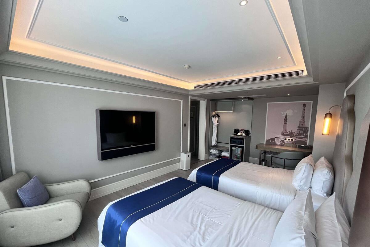 A hotel room with a large flat-screen TV mounted on the wall, a bed with white bedding and blue accents, a seating area with a chair, and a small desk with a mirror and a lamp.