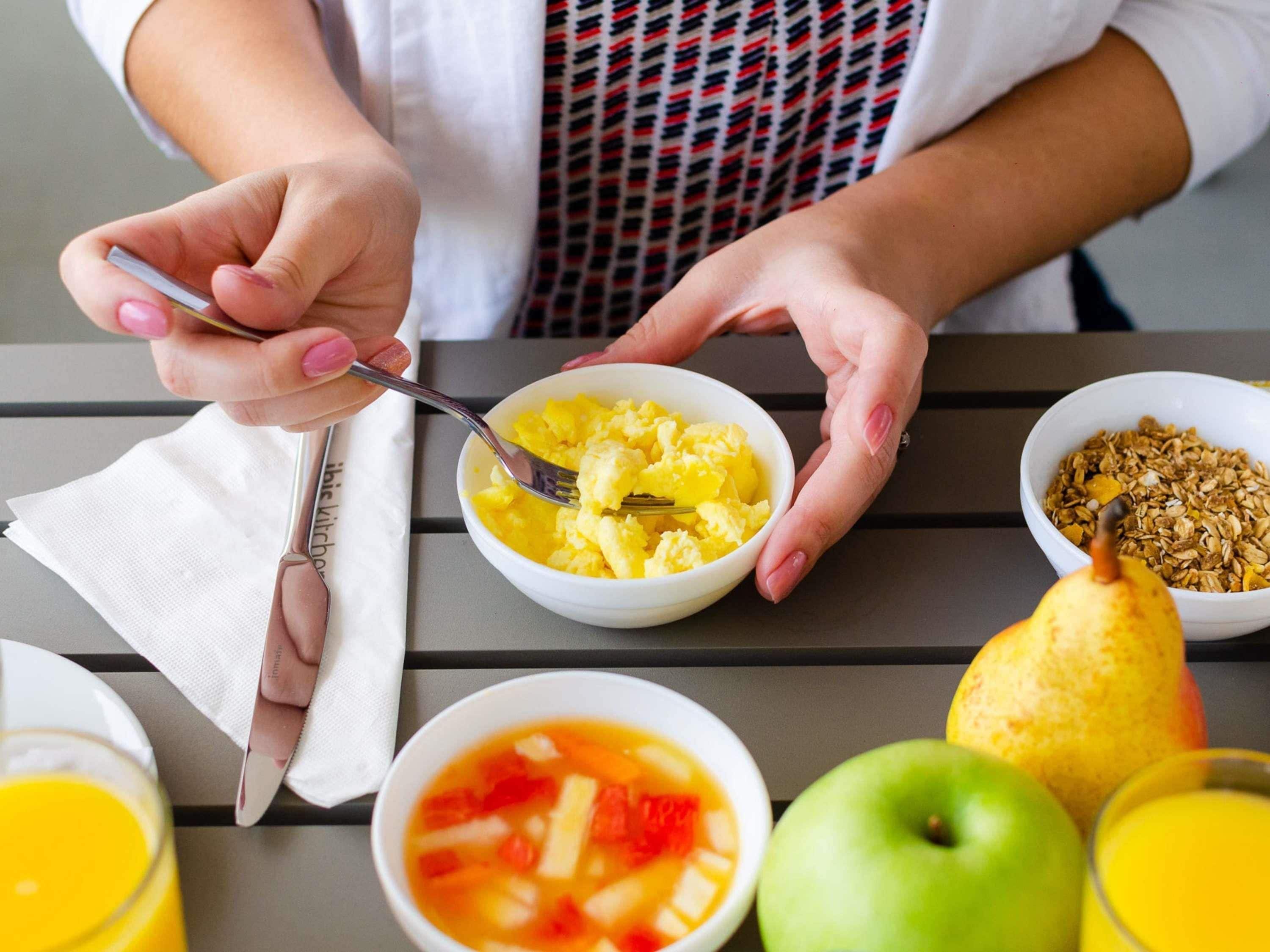 Una persona desayunando huevos revueltos, cereal y fruta en una mesa.