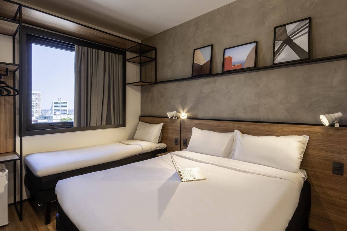 A modern hotel room with a large bed, a smaller bed, a desk, and a window with a city view.