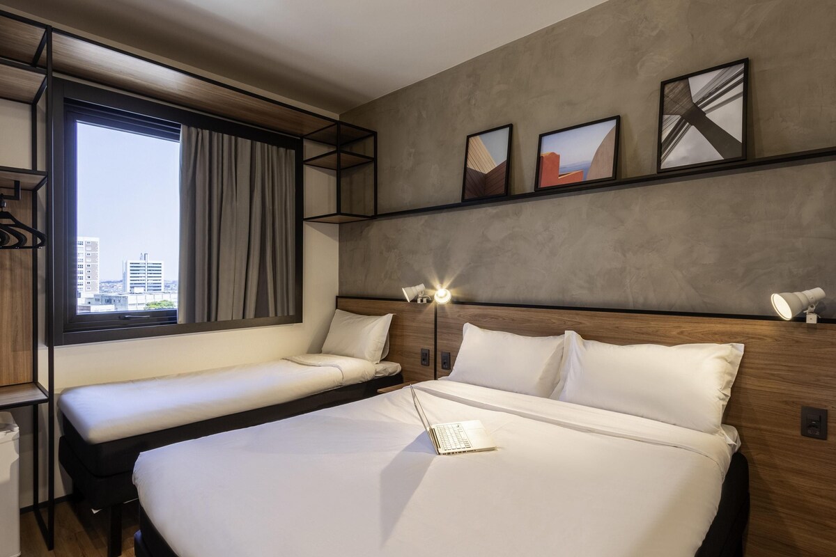 A modern hotel room with a large bed, a smaller bed, a desk, and a window with a city view.