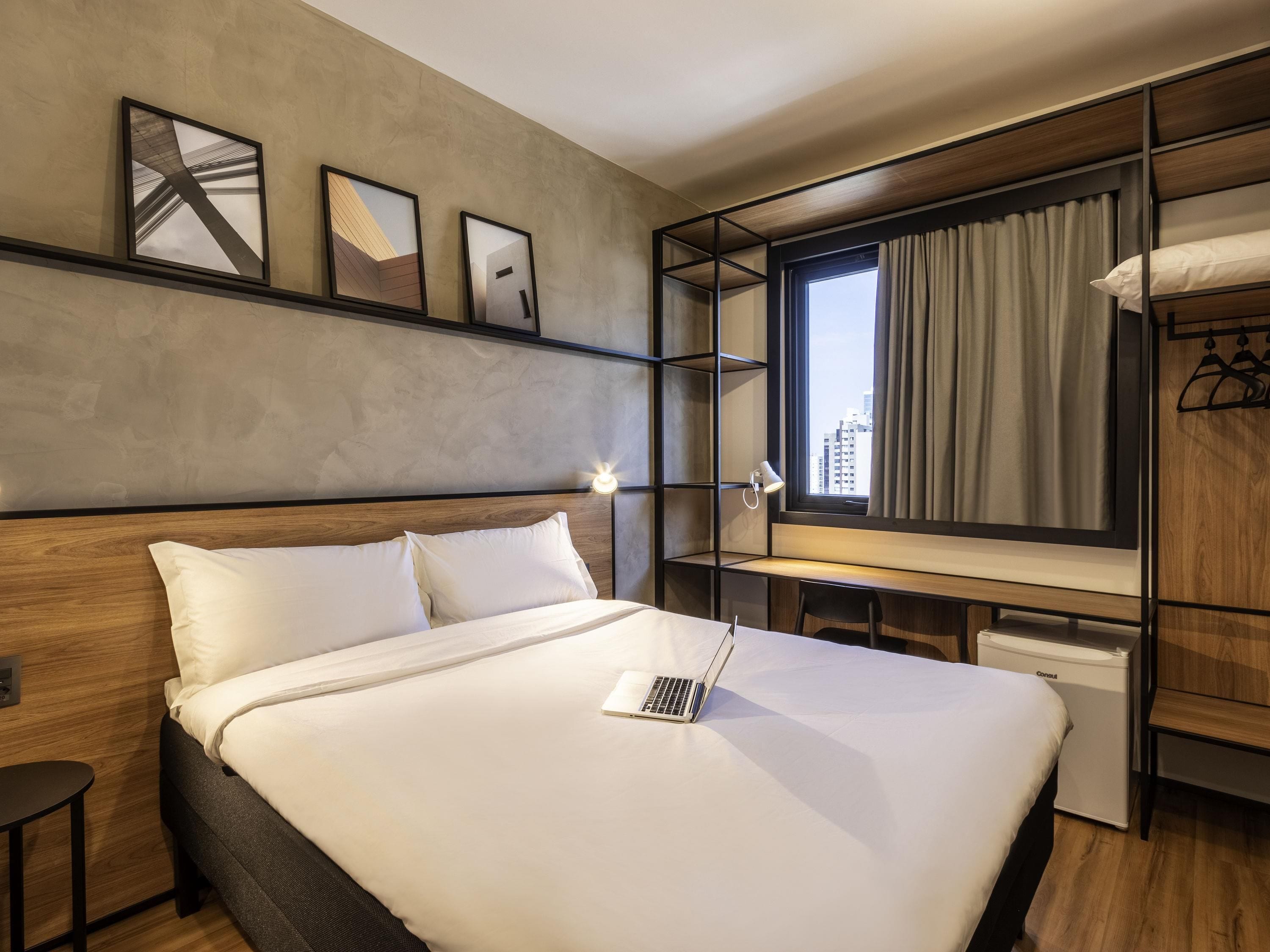 Una habitación de hotel moderna con una cama grande, un escritorio con una computadora portátil, un armario y una ventana con cortinas.