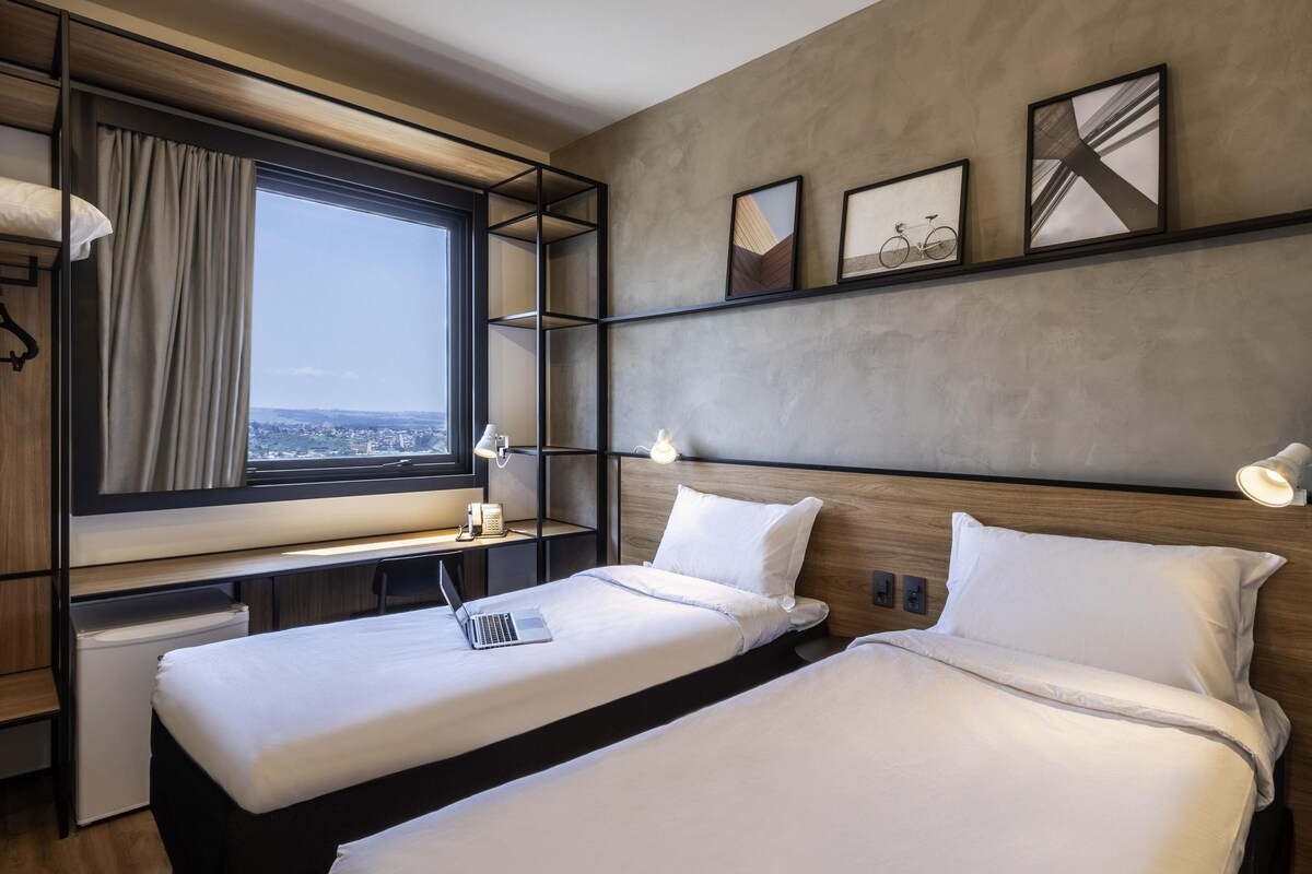 A hotel room with two beds, a desk, a window with a view, and a wall with framed pictures.