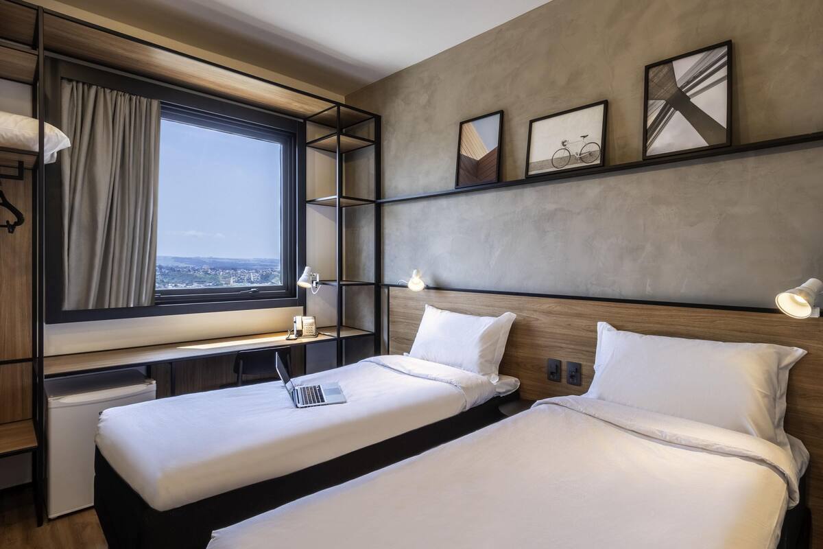 A hotel room with two beds, a desk, a window with a view, and a wall with framed pictures.