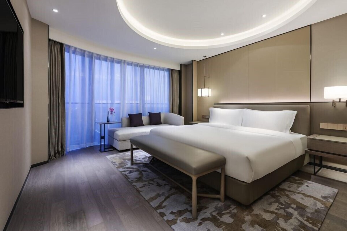 A modern hotel room with a large bed, a bench, a small table, and a large window with curtains.
