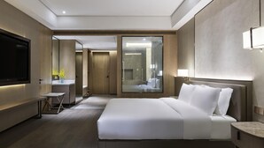 Una habitación de hotel moderna con una cama grande, un televisor de pantalla plana, un escritorio y un baño visible a través de una puerta de cristal.