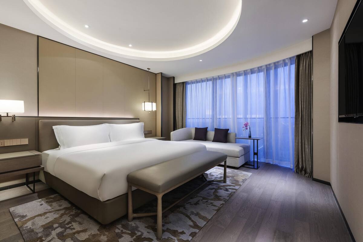 A modern hotel room with a large bed, a sofa, a small table, and a large window with curtains.