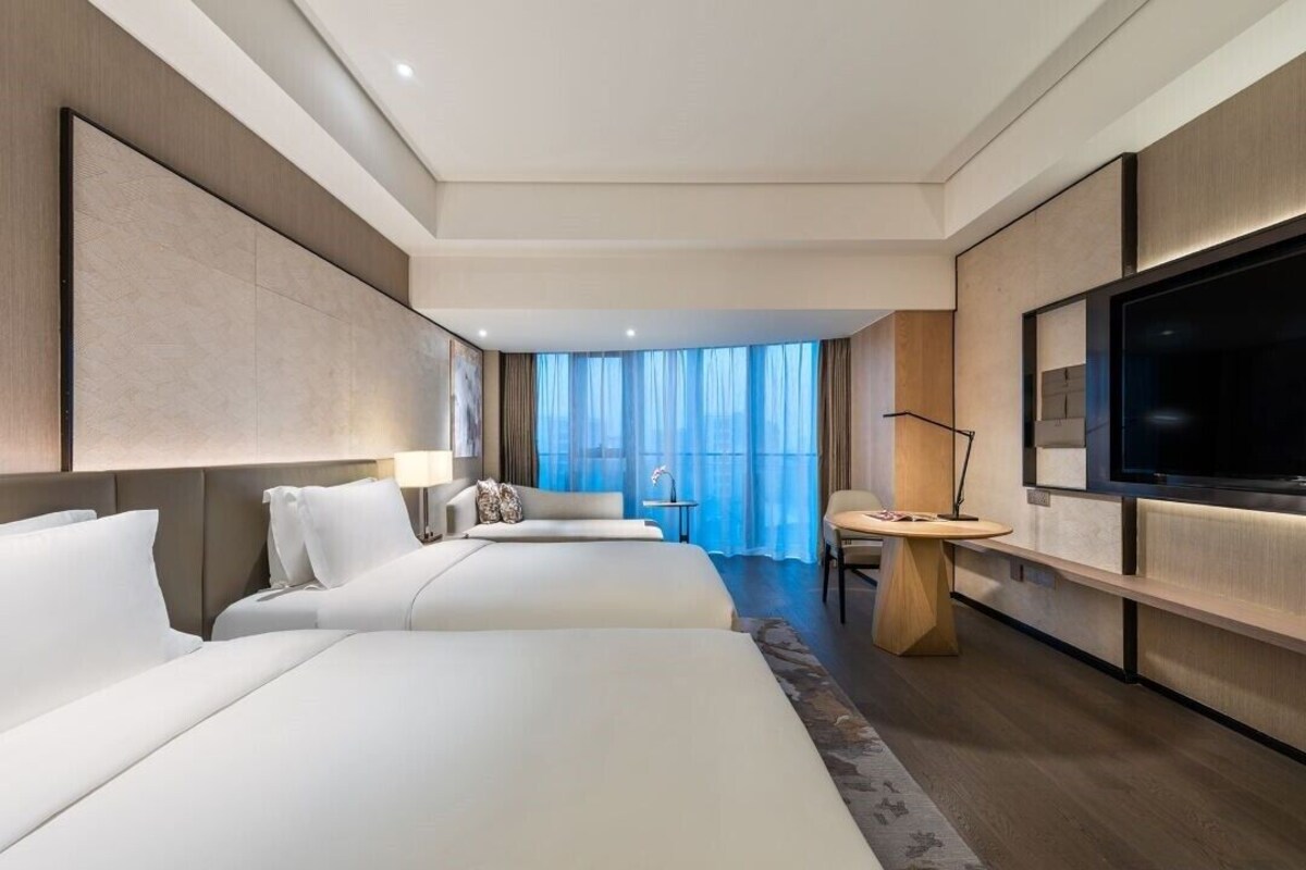 A hotel room with two beds, a TV, a desk, and a view of the city.
