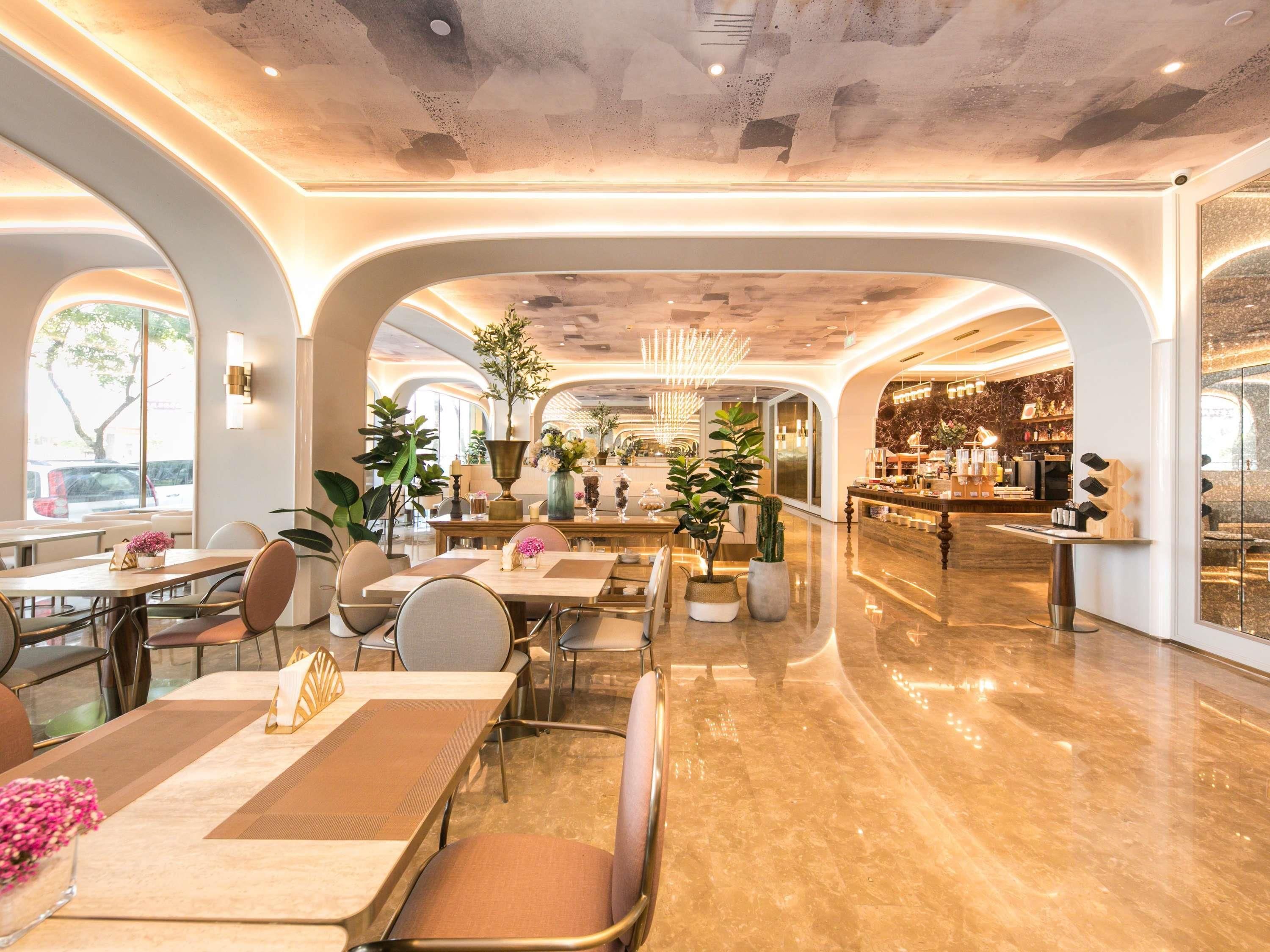 Un amplio vestíbulo de hotel con un bar central, varias zonas de estar y grandes ventanales que dejan entrar luz natural.