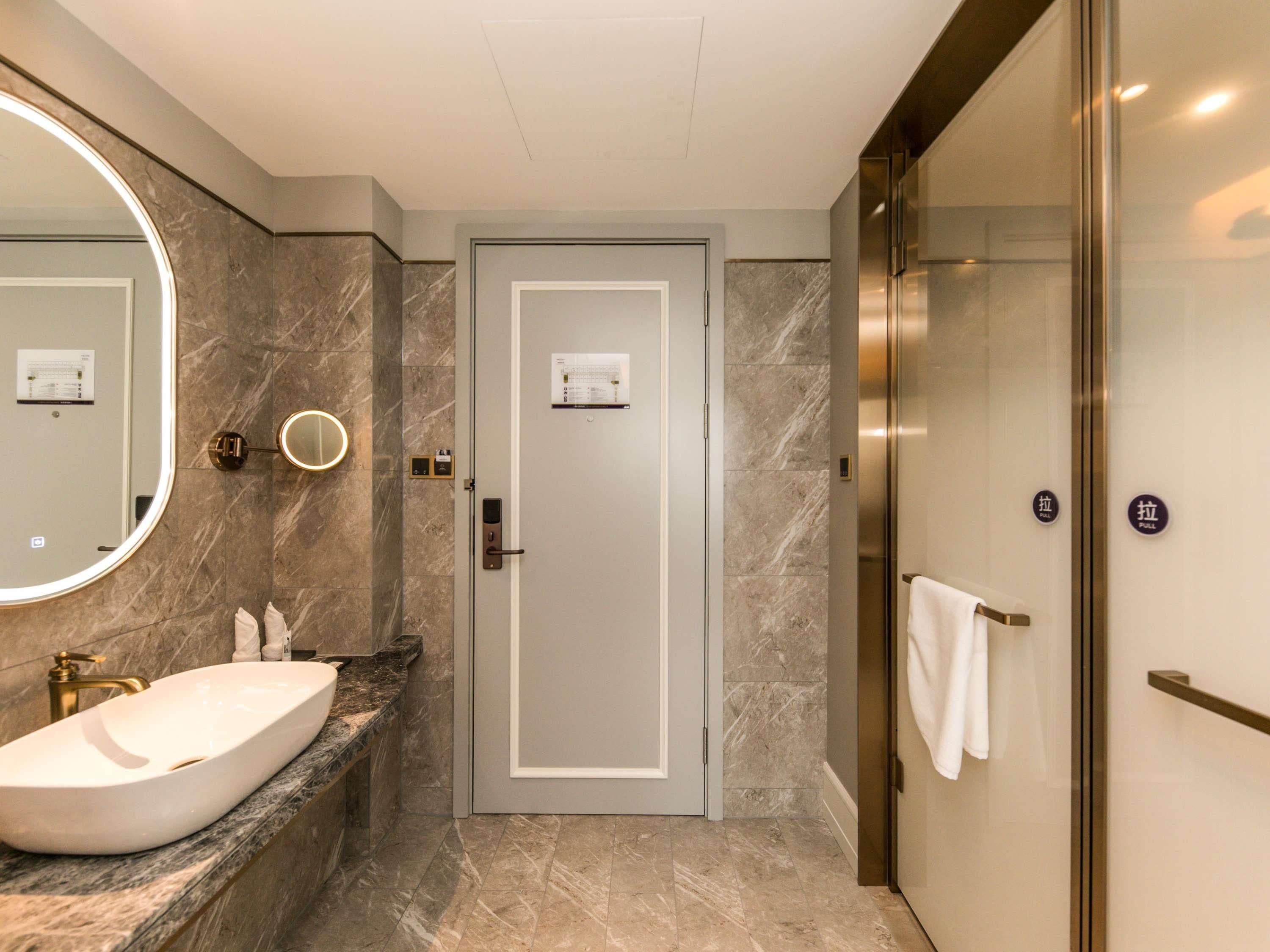 A modern bathroom with a marble sink, a large mirror, a door with a towel rack, and a glass shower enclosure.