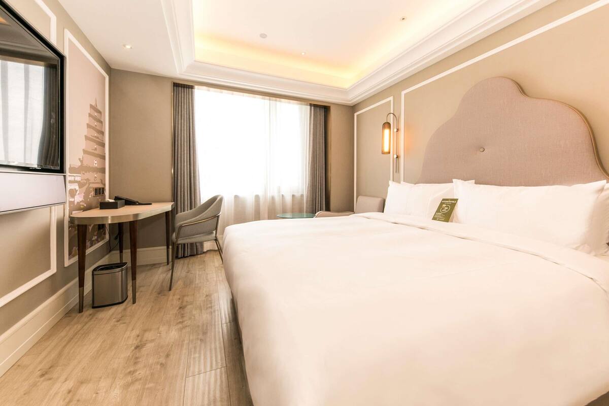 Quarto de hotel com cama grande, escrivaninha, cadeira, televisão e janela com cortinas.