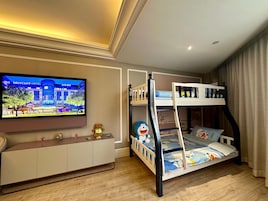 Una cama litera con una colcha de motivos de dibujos animados, un televisor mostrando un anuncio de hotel y un mueble pequeño con figuras decorativas.