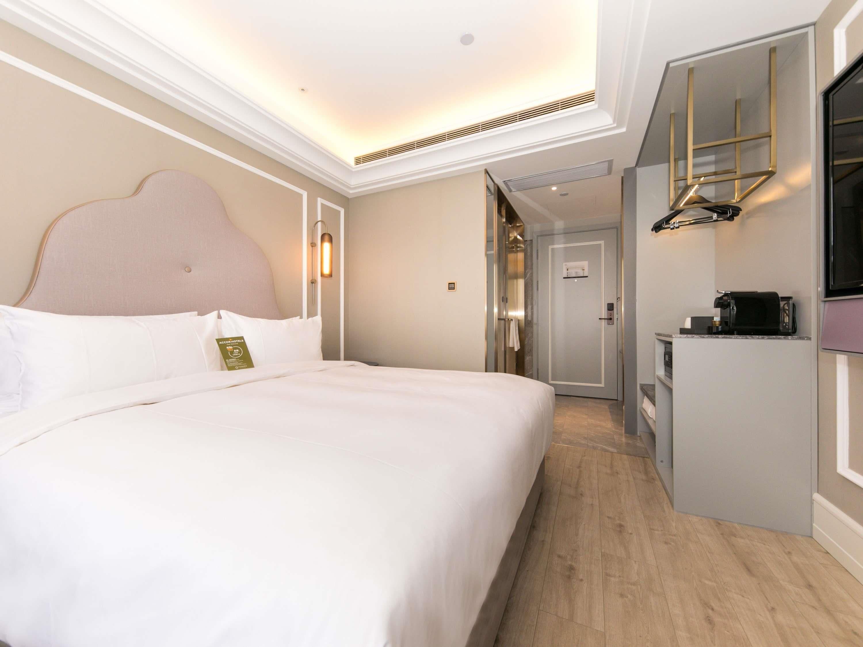 A hotel room with a large bed, a desk with a coffee machine, a mirror, and a door leading to a bathroom.