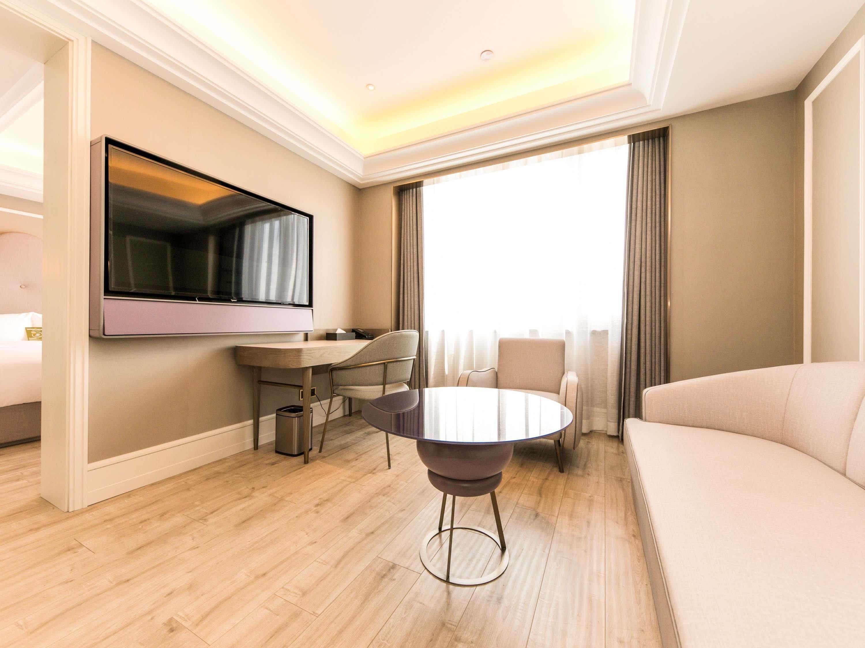 A modern hotel room with a flat-screen TV mounted on the wall, a round dining table with chairs, a sofa, and a large window with curtains.