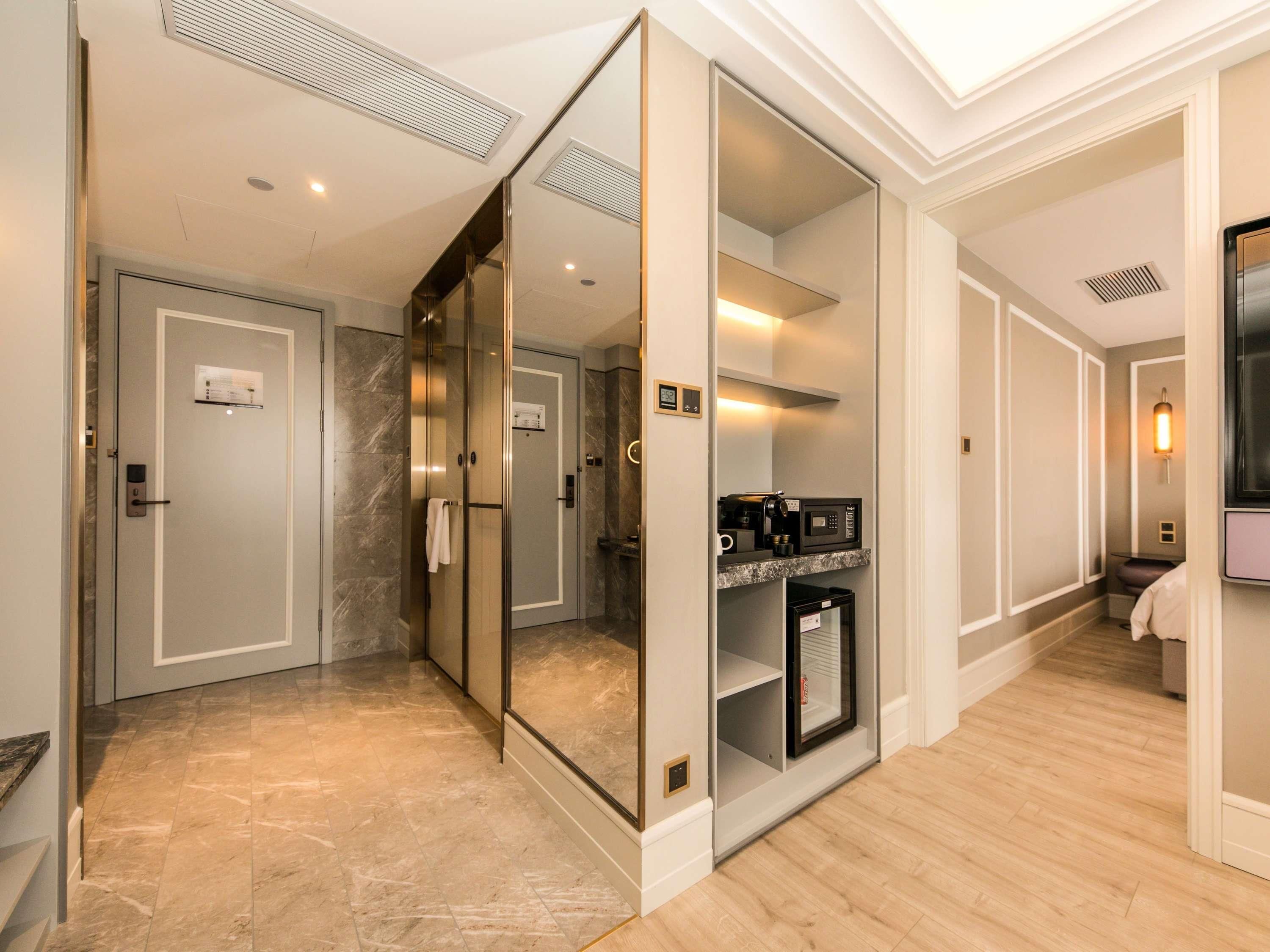 A hotel corridor with a mirrored wall, a glass elevator, a coffee machine, and a microwave.