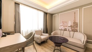 Habitación de hotel moderna con zona de comedor, sofá, mesa de centro y un amplio ventanal con cortinas.