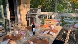 A wooden table set for dining outdoors with a grill, wine glasses, and cutlery.