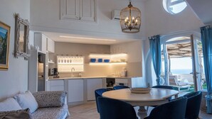 A modern kitchen with white cabinets, a dining area with a round table and blue chairs, and a view of the sea through a large window. - "Villa Costanza" with private pool and jacuzzi sea view - Amalfi Coast (-)