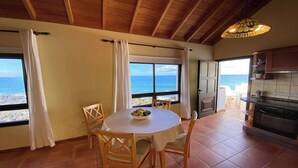 Une pièce avec une table ronde, des chaises, un saladier de fruits, une vue sur la mer, des poutres en bois au plafond et un coin cuisine.