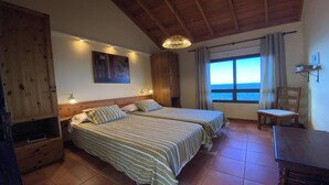 Une chambre avec deux lits, un plafond en bois, une grande fenêtre donnant sur la mer et une chaise près de la fenêtre.