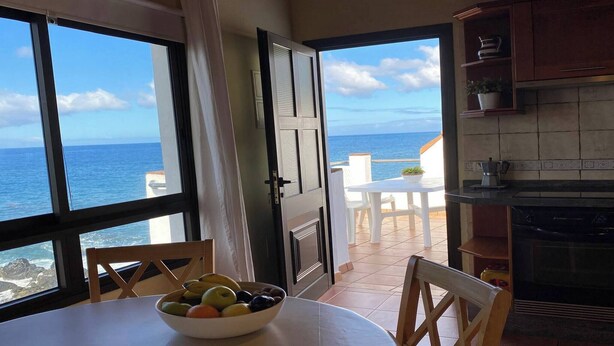 Un coin repas avec vue sur l’océan, un saladier de fruits sur la table et une cuisine équipée d’un lave-vaisselle.