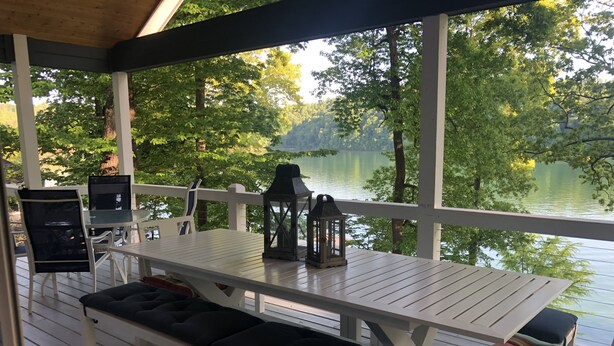 Un espacio de estar techado al aire libre con una mesa y bancos, con vistas a un lago rodeado de árboles.