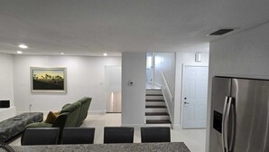 Une cuisine moderne avec un réfrigérateur en acier inoxydable, un coin repas comprenant une table et des chaises, un escalier menant à un deuxième étage, et un tableau représentant un paysage, encadré, accroché au mur.
