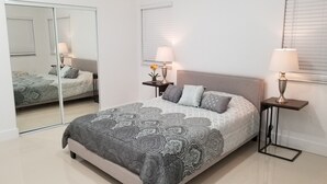 Une chambre avec un grand miroir, un lit avec une literie à motifs, deux tables de chevet avec des lampes et une fenêtre avec des stores.