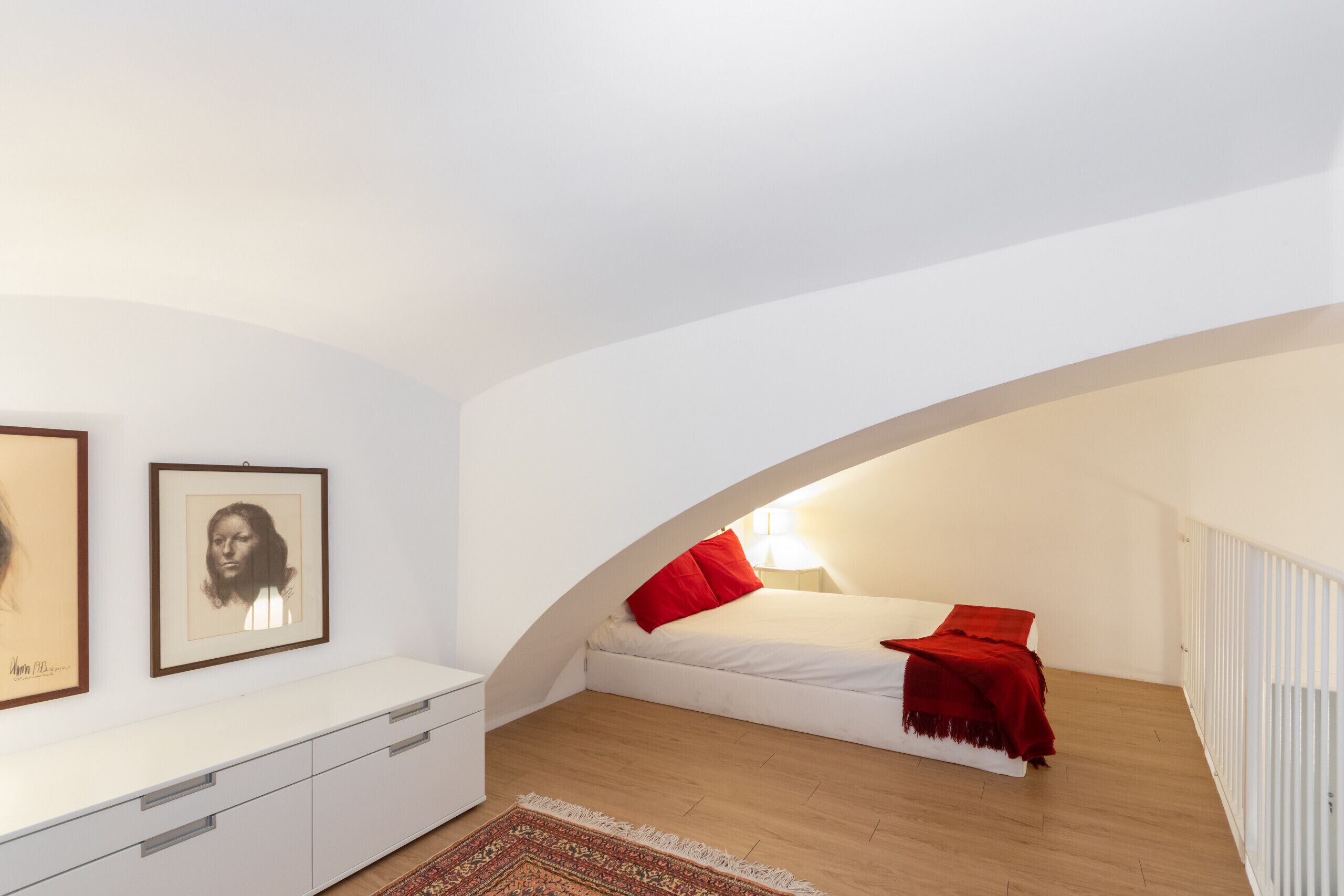 Un dormitorio con una cama blanca, almohadas rojas, un tocador blanco y piso de madera.