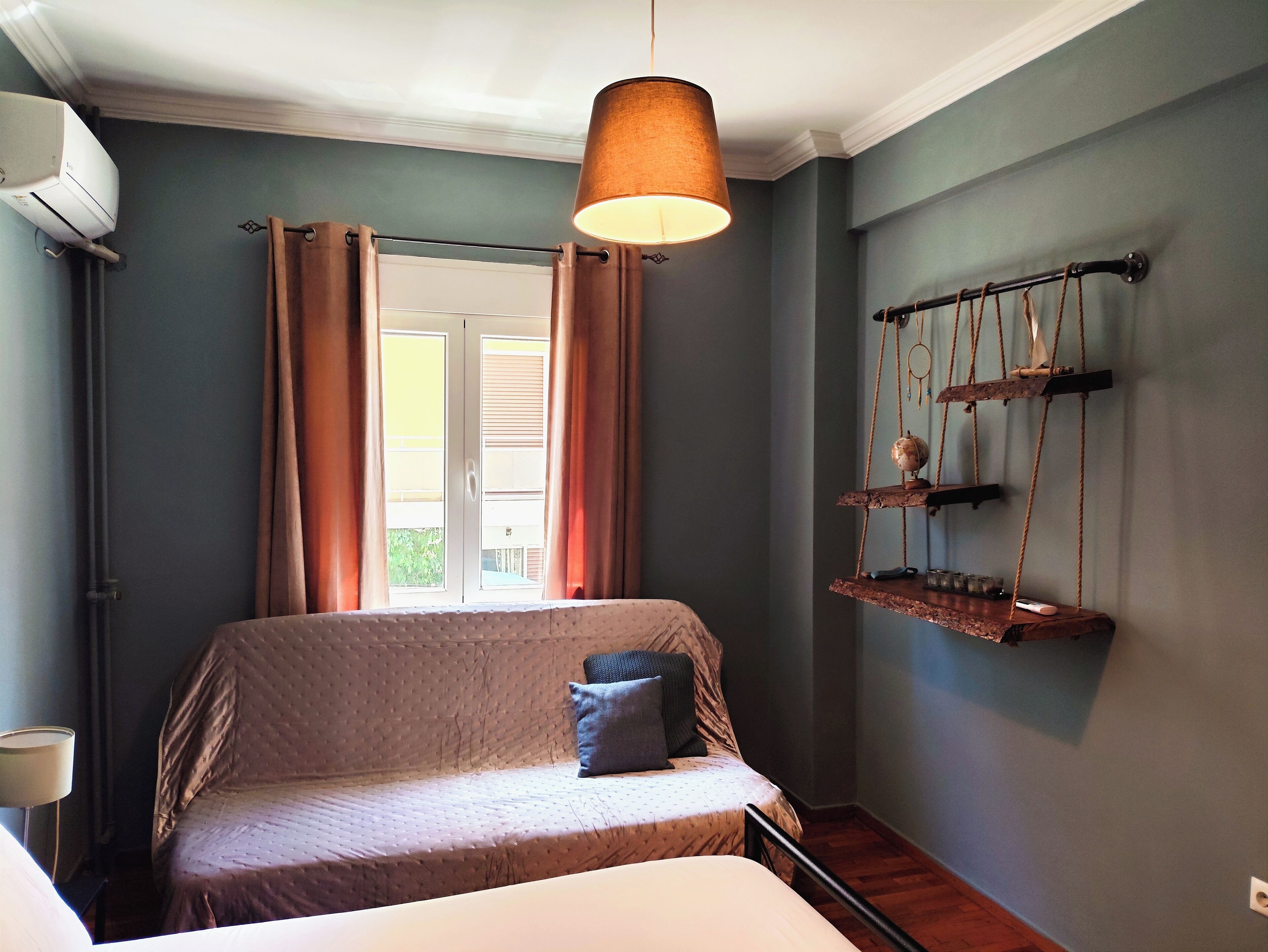 A bedroom with a bed, a hanging light fixture, a window with curtains, and a wall-mounted shelf with items.